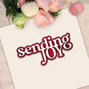 A square cream card with the words 'sending JOY' in white script with a dark red outline, styled with pink roses and green leaves at the top on a light fabric background.