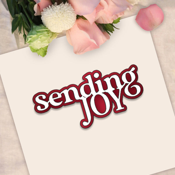 A square cream card with the words 'sending JOY' in white script with a dark red outline, styled with pink roses and green leaves at the top on a light fabric background.