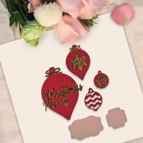 A flat-lay of red Christmas ornament-shaped craft pieces with gold glitter and gold lettering on a white card, styled with pink flowers at the top.