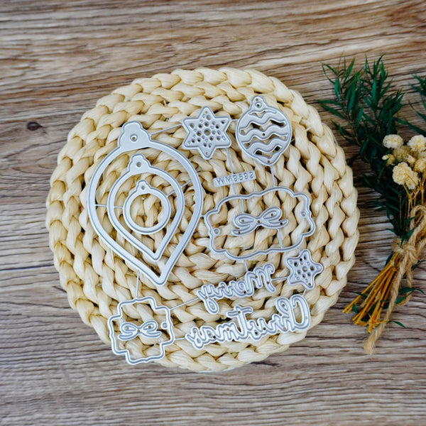 A set of silver metal cookie cutters with Christmas-themed designs, including ornaments, stars, and holiday phrases, arranged on a round woven straw mat over a wooden surface.