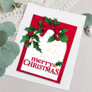 A handmade Christmas greeting card with a red and white design, decorated with green holly leaves, a red bow, and gold accents, reading 'merry CHRISTMAS'. The card is styled on a textured white surface with eucalyptus leaves.