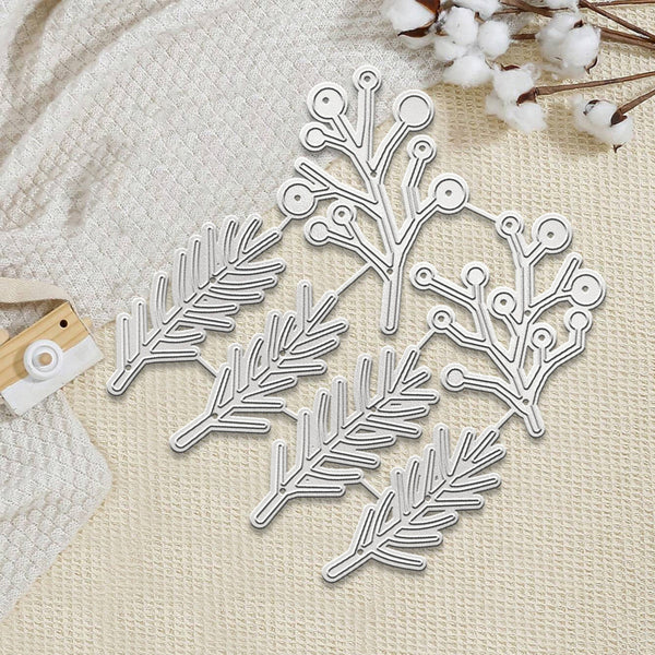 A set of silver metal die-cut embellishments shaped like pine branches and leaves, arranged on a textured cream fabric with cotton stems as decorative props.