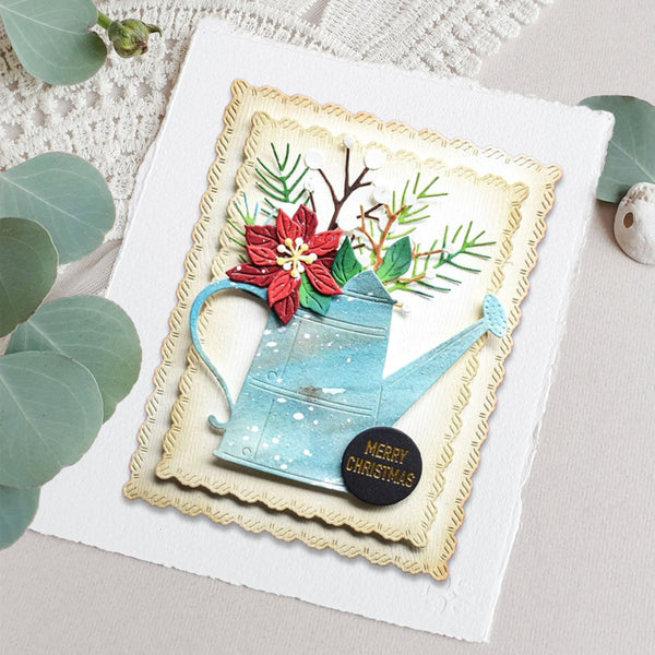 A handmade Christmas greeting card featuring a light blue watering can with a red poinsettia and green pine branches, layered over a cream scalloped border and placed on a white cardstock. The card is styled with eucalyptus leaves and a small wooden bead on a neutral fabric background.