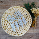 Four silver metal leaf-shaped nail art stamps arranged on a round woven straw mat, with dried flowers and greenery on a rustic wooden surface.