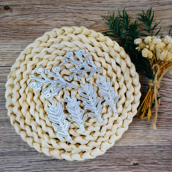 Four silver metal leaf-shaped nail art stamps arranged on a round woven straw mat, with dried flowers and greenery on a rustic wooden surface.