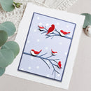 A square handmade card featuring layered red and white birds perched on dark blue branches against a pale lavender background with white polka dots, styled on a textured white surface with green leaves around it.