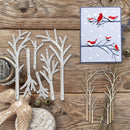 A flat-lay of winter-themed crafting supplies on a rustic wooden surface, featuring a set of white metal tree dies, finished twig-style tree cutouts in natural wood, a small handmade card with red birds, and decorative props including a ceramic bird figurine and jingle bells.