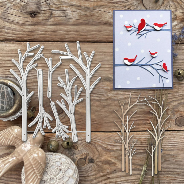 A flat-lay of winter-themed crafting supplies on a rustic wooden surface, featuring a set of white metal tree dies, finished twig-style tree cutouts in natural wood, a small handmade card with red birds, and decorative props including a ceramic bird figurine and jingle bells.