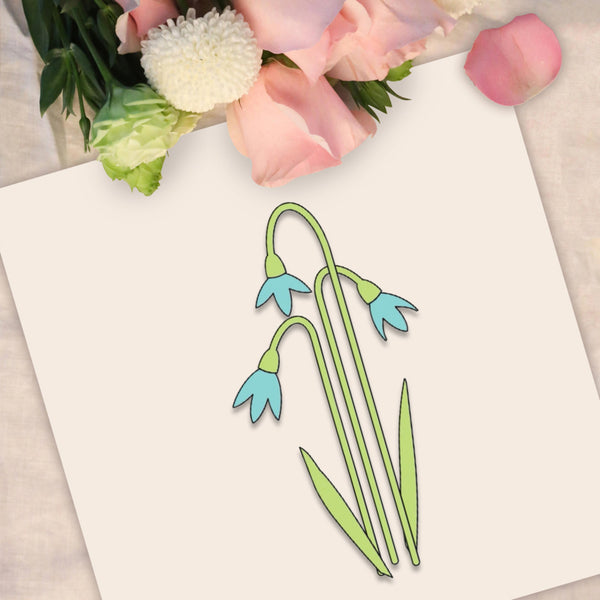 A decorative flat-lay showing a white card with a simple line drawing of three snowflake-like flowers in light green and light blue, surrounded by pink and white flowers on a light fabric surface.