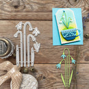 A flat-lay of a crafting set featuring white metal die-cut flowers on a rustic wooden background, alongside a finished layered card showing a blue cup and white lily of the valley flowers on a blue and yellow background.