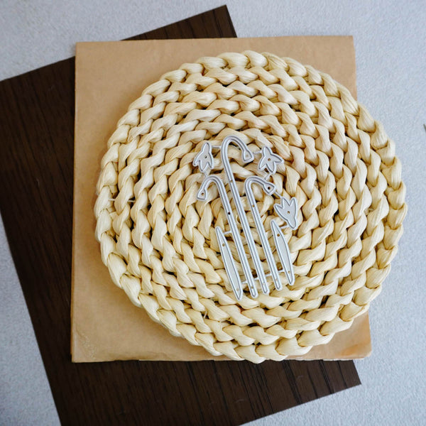 A round woven straw placemat in a light cream color, displayed on a square tan paper background with a silver metal monogram template placed in the center.