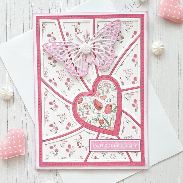 A handmade pink and white anniversary card with a butterfly and heart design, displayed on a white wooden surface with decorative pink and white accessories.