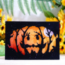A rectangular black card or plaque featuring a watercolor-style jack-o'-lantern face with orange and red tones, overlaid with black tree silhouettes and a pale moon, displayed on a light surface with blurred yellow flowers and green leaves in the background.