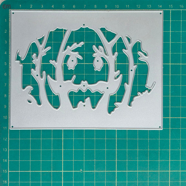 A metal cutting die with a spooky jack-o'-lantern face and tree silhouette cutout, displayed on a green cutting mat with grid lines.