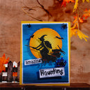 A handmade Halloween greeting card featuring a witch riding a broomstick in front of a large yellow moon, with blue striped background elements and text reading 'amazing Haunting'. The card is displayed on a wooden surface against a rustic brown wall with autumn leaves.