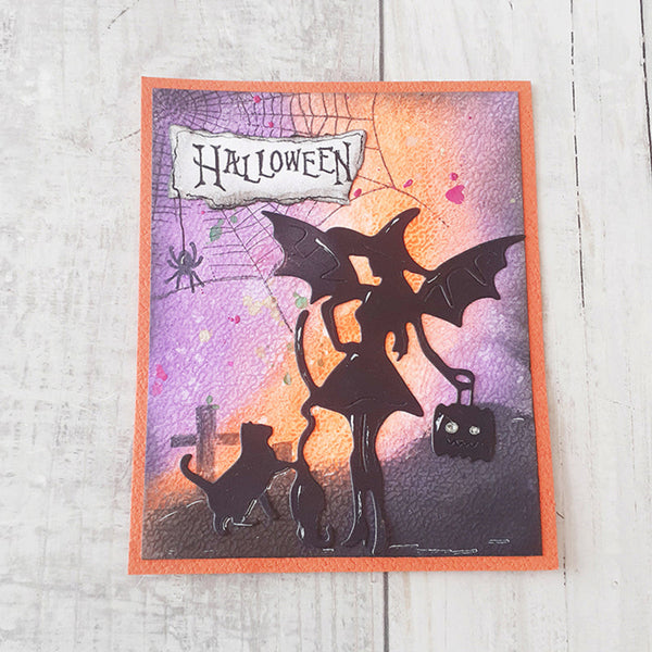 A square Halloween-themed card featuring a black silhouette of a witch with a broom, a cat, a spider, and a small black jack-o'-lantern, set against a purple and orange sunset background with a torn-paper 'HALLOWEEN' banner. The card has an orange border and is photographed on a light wood surface.