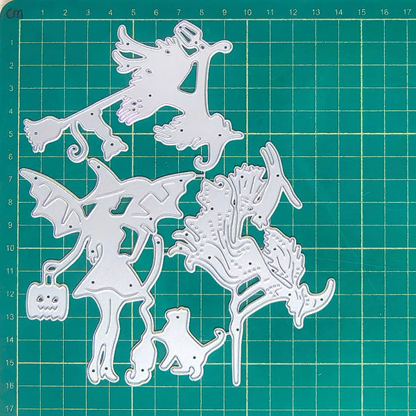 A set of white metal die-cut craft shapes featuring whimsical fairy, dragon, cat, and pumpkin designs, arranged on a green cutting mat with grid lines and centimeter measurements.