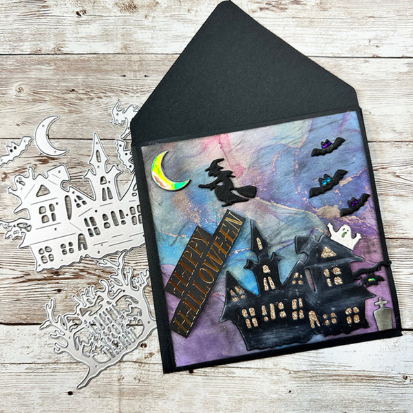 A Halloween-themed handmade greeting card with a black envelope, featuring a haunted house, witch on a broomstick, flying bats, a crescent moon, and ghost cutouts. Silver die-cut embellishments are scattered on the rustic wood background.