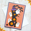 A Halloween-themed layered card featuring the word 'BOO' in black with orange cutouts, surrounded by white ghosts, black bats, a purple witch hat, and an orange polka-dot background, displayed on a white envelope over a light wood surface.