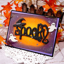 A handmade Halloween greeting card with 'spooky' in glittery black letters, decorated with white ghost cutouts, a witch hat, and a broom, displayed on a white lace doily with autumn leaves and a picket fence backdrop.