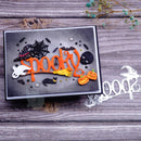 A rectangular Halloween-themed craft or card featuring raised orange 'spooky' lettering with black bats, skulls, a witch hat, a yellow broom, and small ghost and pumpkin details on a gray background, photographed on a rustic wood surface with a metal die cutout nearby.