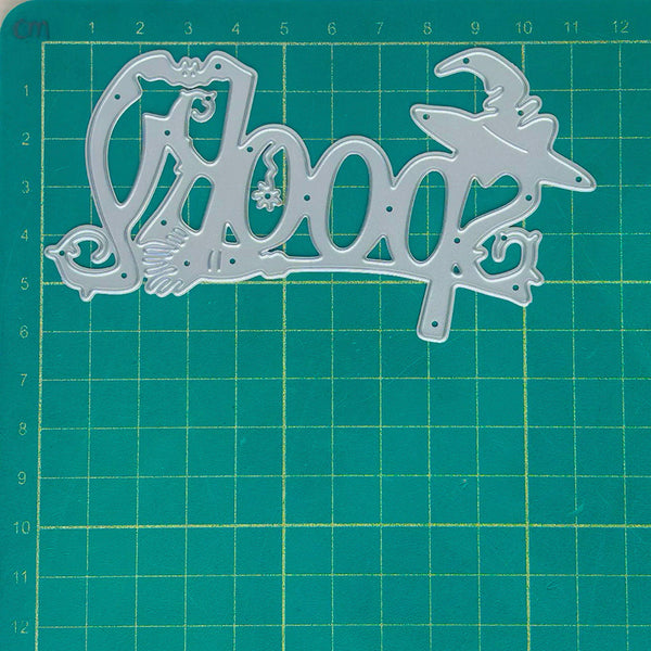 A silver metal die-cut craft stencil with the word 'hoop' in decorative script and a witch hat, placed on a green cutting mat with a yellow grid.