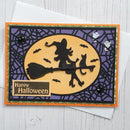 A Halloween-themed layered card featuring a witch riding a broomstick with a cat on her head, set against a purple spiderweb pattern and a yellow moon. The card includes a 'Happy Halloween' label and is displayed on top of a white envelope over a white wood background.