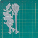A silver metal cutting die set featuring a witch and cat silhouette and a broom, displayed on a green self-healing cutting mat with grid lines and measurements.