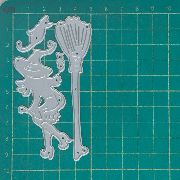 A silver metal cutting die set featuring a witch and cat silhouette and a broom, displayed on a green self-healing cutting mat with grid lines and measurements.