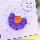 A close-up of a handmade greeting card featuring a purple paper flower with a layered orange center, embossed white swirls, and a circular 'Thanks so Much' message in the top right.