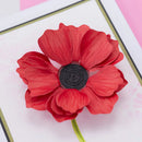A red paper flower embellishment with layered petals and a black rolled center, attached to a green stem, photographed close up on a white card with pink and gold border details.