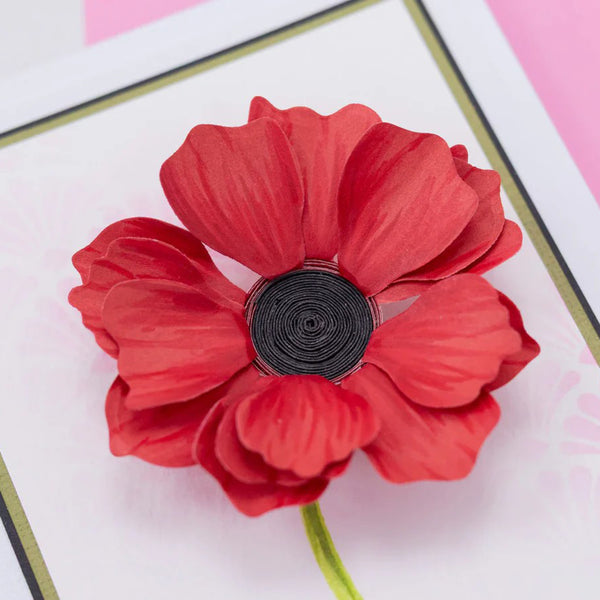 A red paper flower embellishment with layered petals and a black rolled center, attached to a green stem, photographed close up on a white card with pink and gold border details.