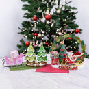 A festive Christmas tabletop display with miniature green trees, a pink sleigh with a gift box, a wreath, and a red sleigh with candy and a 'Merry Christmas' sign, set in front of a blurred decorated Christmas tree.