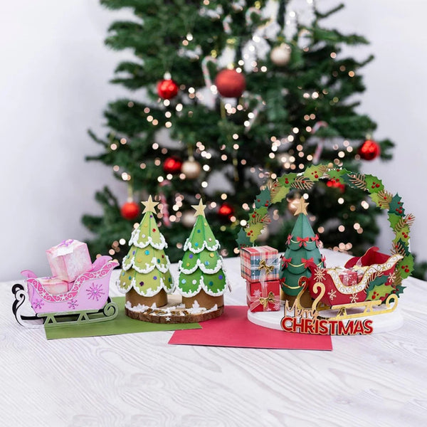 A festive Christmas tabletop display with miniature green trees, a pink sleigh with a gift box, a wreath, and a red sleigh with candy and a 'Merry Christmas' sign, set in front of a blurred decorated Christmas tree.