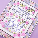 A close-up of a decorative layered card or craft plaque with the phrase “SISTER YOU’RE THE Best” in purple lettering, surrounded by pink flowers and gold leaf-like floral accents on a floral lavender background.