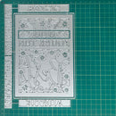 A silver metal die-cutting or stamping template with floral and script motifs, shown on a green cutting mat with measurement markings.