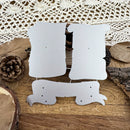 Three white cardboard or paper cutout shapes with pre-drilled holes, arranged on a rustic wooden surface with dried leaves, a pinecone, bark, and a lace cloth in the background.