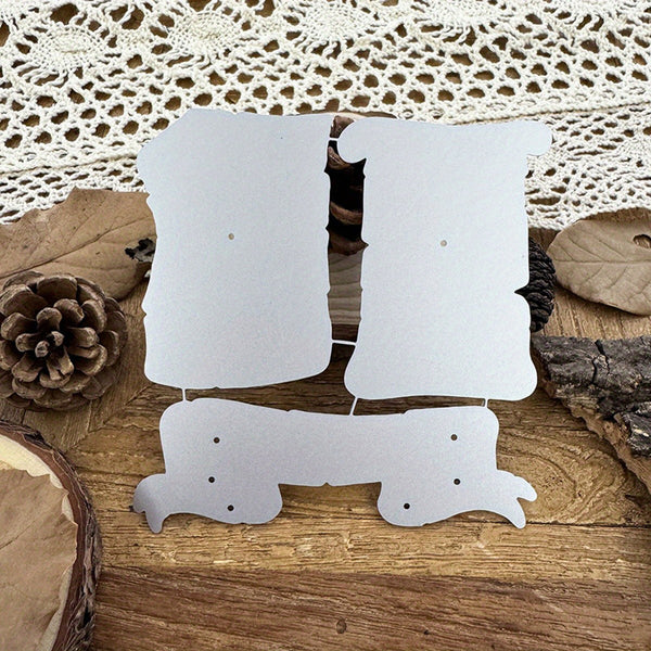 Three white cardboard or paper cutout shapes with pre-drilled holes, arranged on a rustic wooden surface with dried leaves, a pinecone, bark, and a lace cloth in the background.