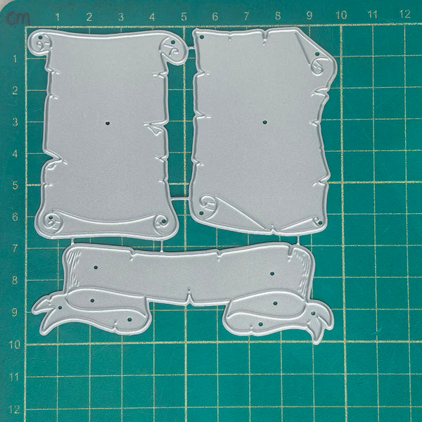 Three clear plastic embossing plates with scroll and banner designs, laid out on a green cutting mat with grid lines.