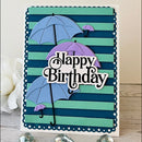 A handmade square birthday card with layered paper umbrellas in light blue and lavender, set against teal and navy horizontal stripes. The card features a scalloped dark blue border and a central die-cut white label reading 'Happy Birthday' in black script.