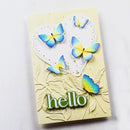 A handmade greeting card with a pale yellow background, featuring embossed leaf details, a white heart-shaped cutout, and four blue and yellow butterflies. The card is decorated with the word 'hello' in green 3D letters and is photographed on a white textured surface.