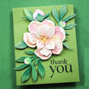A handmade paper card featuring layered pink and white flowers with green leaves on a green background, with the words 'thank you' printed in black script.