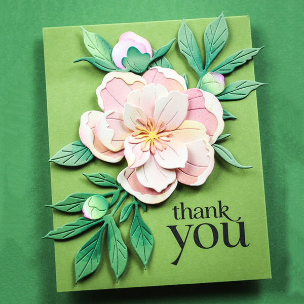 A handmade paper card featuring layered pink and white flowers with green leaves on a green background, with the words 'thank you' printed in black script.