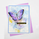 A handmade birthday card featuring a purple and lavender butterfly on a watercolor background with blue and yellow washes, layered over a purple envelope. The card has a blue border and a small black banner reading 'happy birthday' in gold lettering.