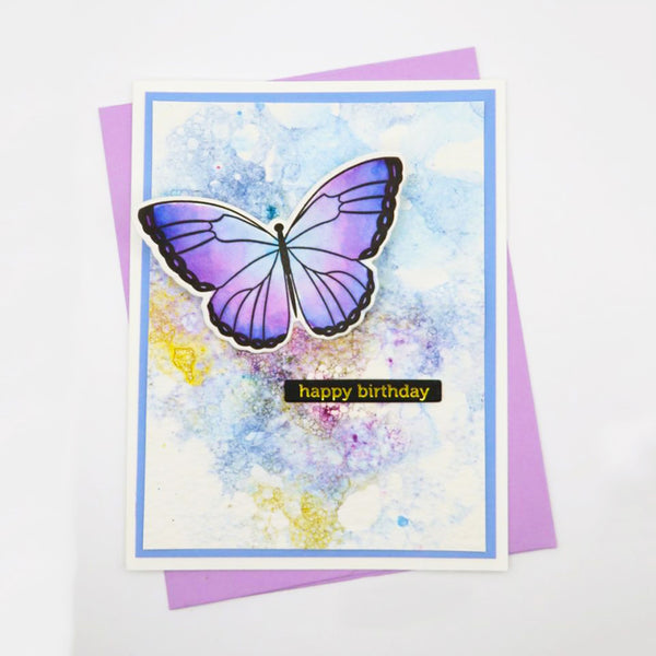 A handmade birthday card featuring a purple and lavender butterfly on a watercolor background with blue and yellow washes, layered over a purple envelope. The card has a blue border and a small black banner reading 'happy birthday' in gold lettering.