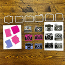 A flat lay of craft dies and sticker sheets featuring retro camera and photo frame designs on a rustic wooden background. The set includes six white metal camera-shaped dies, a sheet of pink and blue paper inserts, and two sheets of stickers showing various camera illustrations in black, gray, pink, and blue.