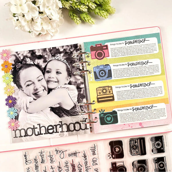 An open ring-bound scrapbook or journaling planner page displayed on a light background with flowers and a decorative sheet of camera-themed stickers below. The page includes a black-and-white photo of a woman and child, colorful camera illustrations, handwritten text, and the word 'motherhood' at the bottom.