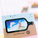 A handmade greeting card featuring a bottle-shaped cutout with a lighthouse, dolphin, and blue sky scene, placed on a kraft paper base with decorative anchor, ship wheel, and the words 'Just a little message'.