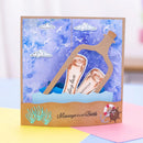 A handmade pop-up greeting card featuring a watercolor blue ocean scene with clouds, a brown cardboard bottle, rolled paper messages reading 'ding love' and 'message in a bottle,' and a sandy shore with small decorative sea-themed stickers.