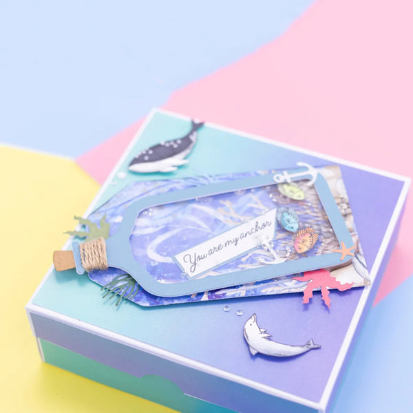 A decorative gift box with a layered pastel design in blue, purple, and teal, featuring a bottle-shaped clear window containing blue glitter and a card that reads 'You are my anchor,' plus small sea-creature embellishments.